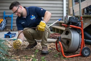 Plumber using a drain snake to clear a sewer clog outdoors, wearing gloves and working near plumbing equipment, emphasizing professional drain cleaning services.