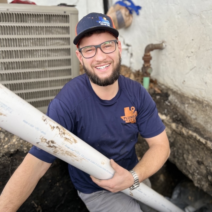 Man smiling while holding a sewer pipe, wearing a branded shirt, in a trench indicating sewer line repair services in Fort Worth.